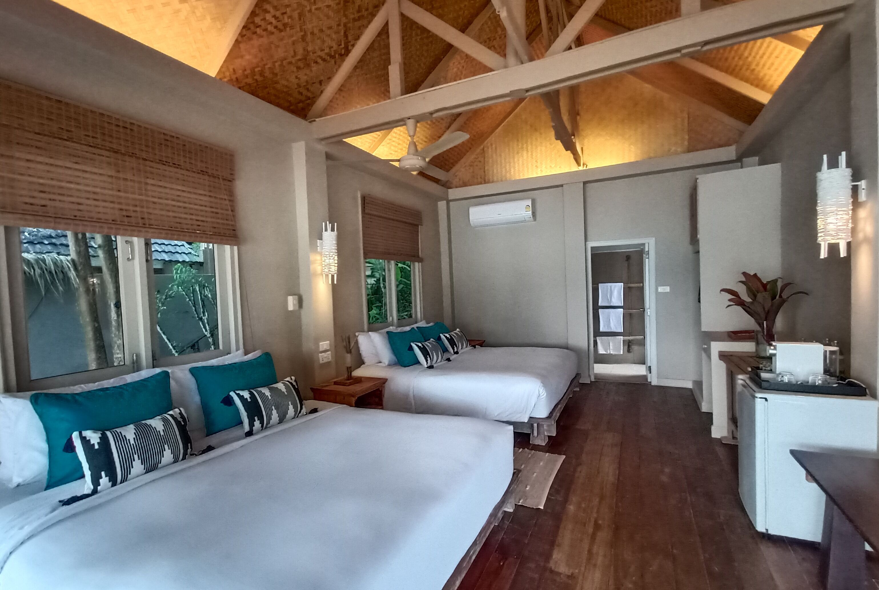 A hotel room with two beds, a ceiling fan, a small kitchen area, and a view of the outdoors.