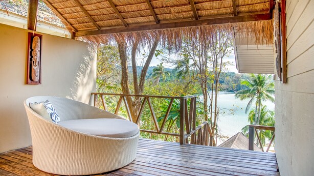 A wicker lounge chair on a wooden deck with a view of a tropical landscape and water.
