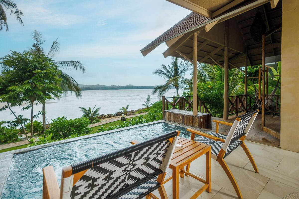 Un espace aménagé au bord de la piscine, avec vue sur la mer, doté de mobilier en bois et de plantes tropicales.