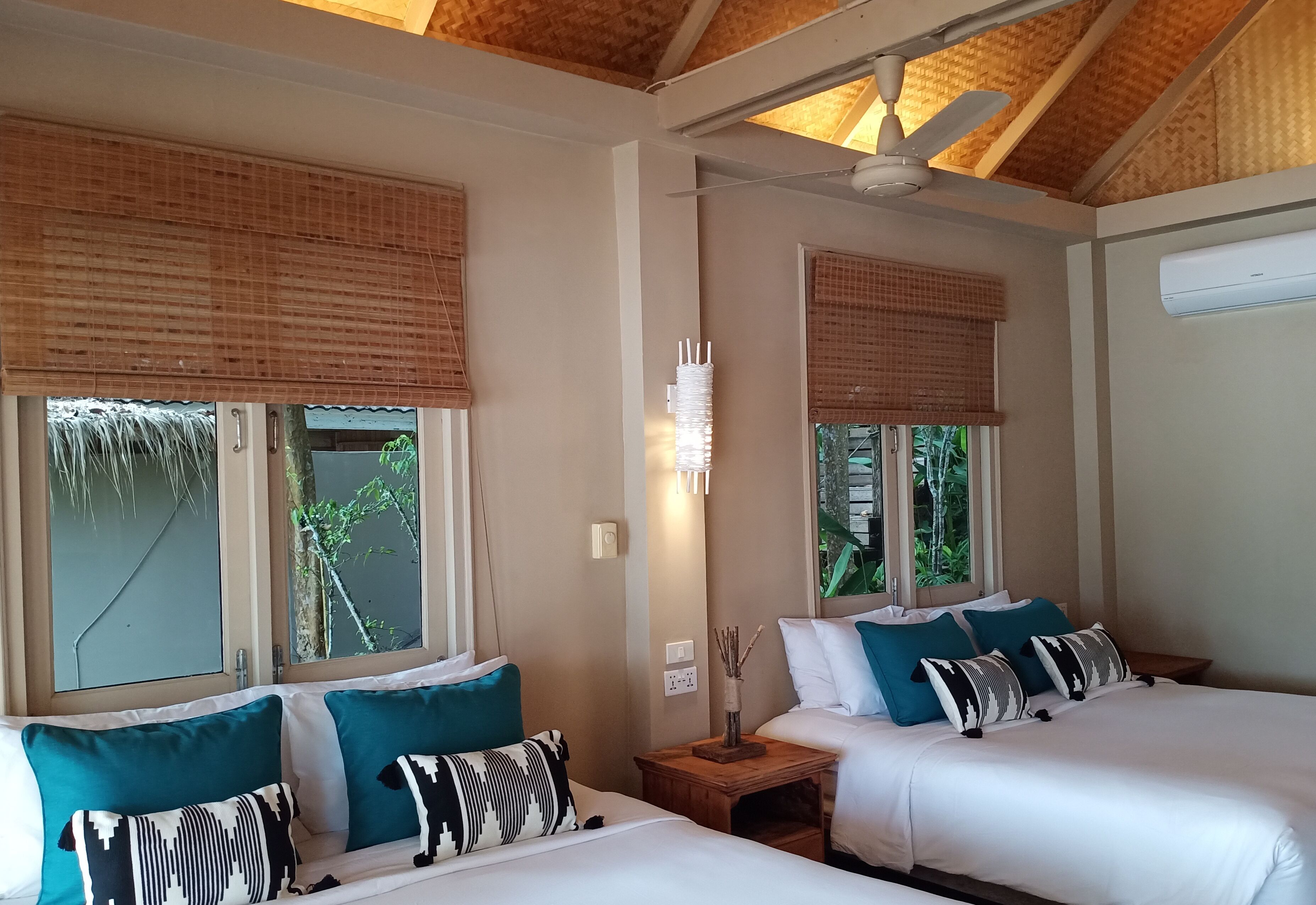 Ein Zimmer mit zwei Betten, jeweils mit wei ßer Bettwäsche und türkisfarbenen Kissen. Holz-Nachttische mit Lampen und eine Wandleuchte sind vorhanden. Große Fenster mit Bambusrollos und eine Klimaanlage an der Wand sind zu sehen.