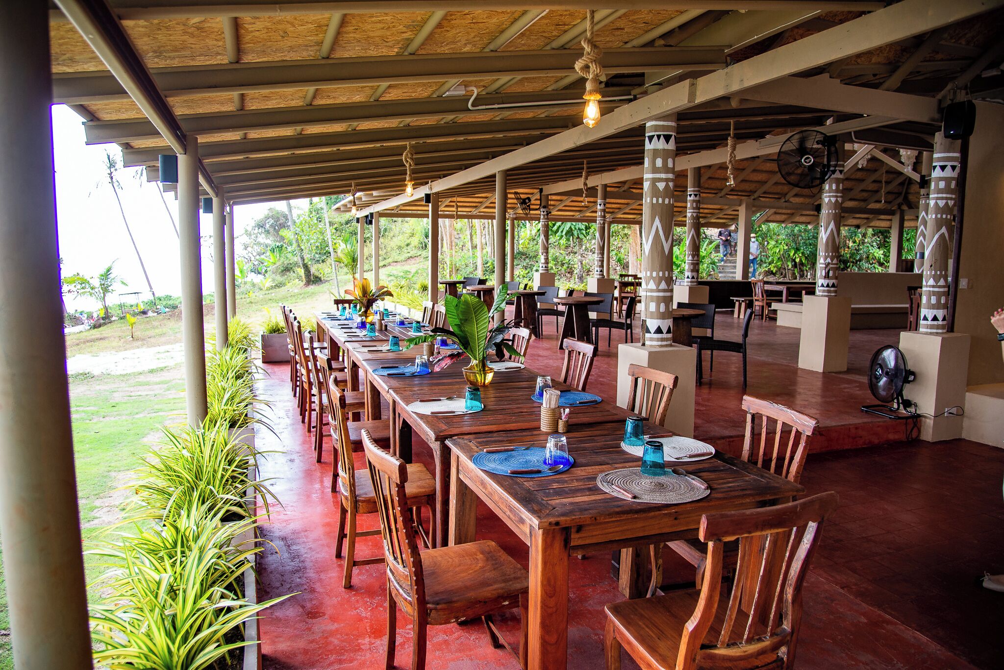 An outdoor dining area with wooden tables and chairs, a covered roof, and a view of the surrounding greenery.