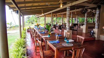 An outdoor dining area with wooden tables and chairs, a covered roof, and a view of the surrounding greenery.
