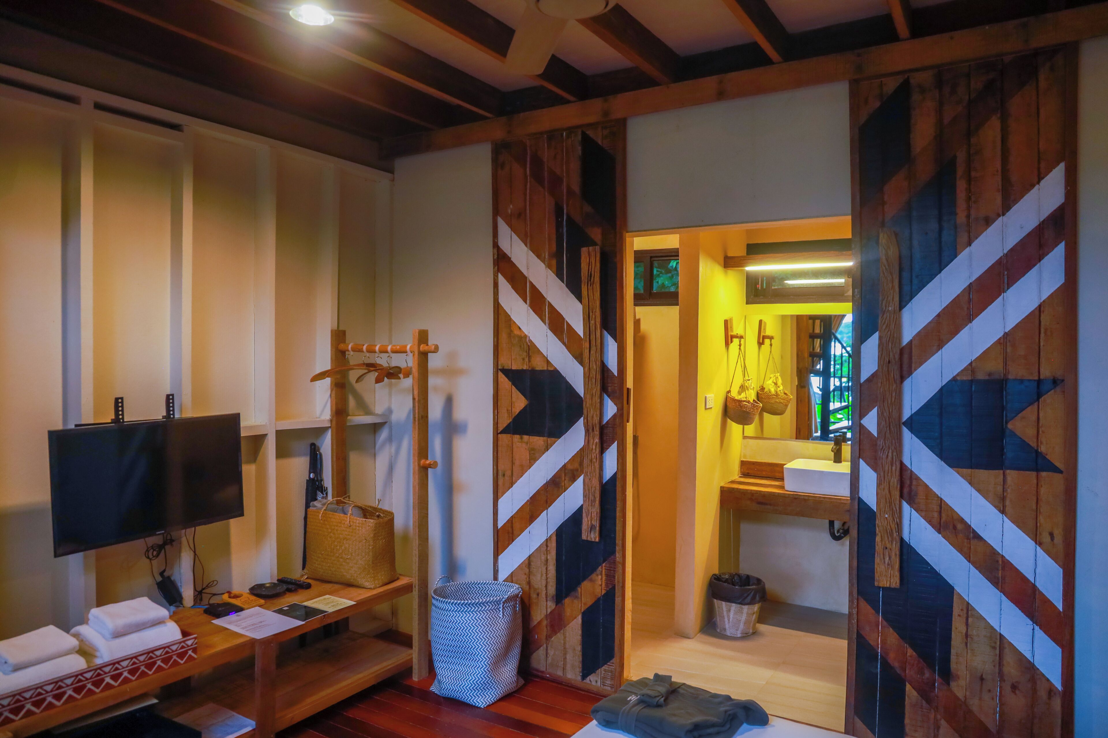 A room with a television, a wooden bench, a basket, a waste bin, and a door with a geometric pattern.