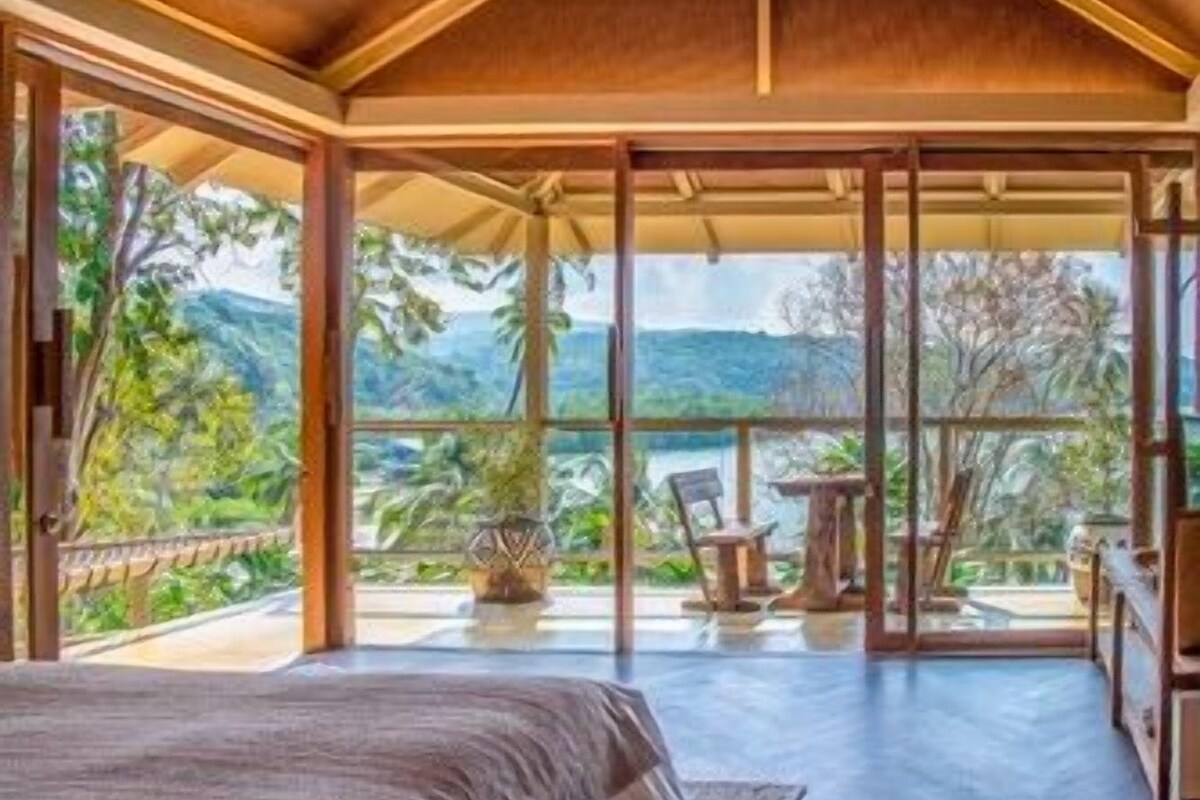 A bedroom with a large bed, a balcony with a view of greenery and hills, and a dining area with chairs and a table.