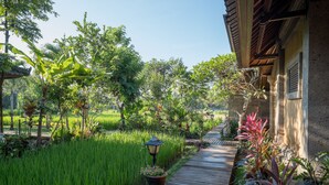 Un sentier en bois mène à un bâtiment à l’architecture traditionnelle, entouré d’une végétation luxuriante et d’un jardin bien entretenu.