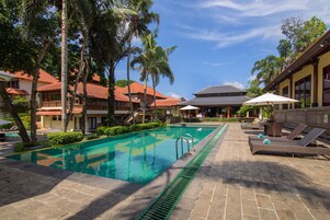 A swimming pool with lounge chairs and umbrellas, surrounded by palm trees and buildings with red roofs.