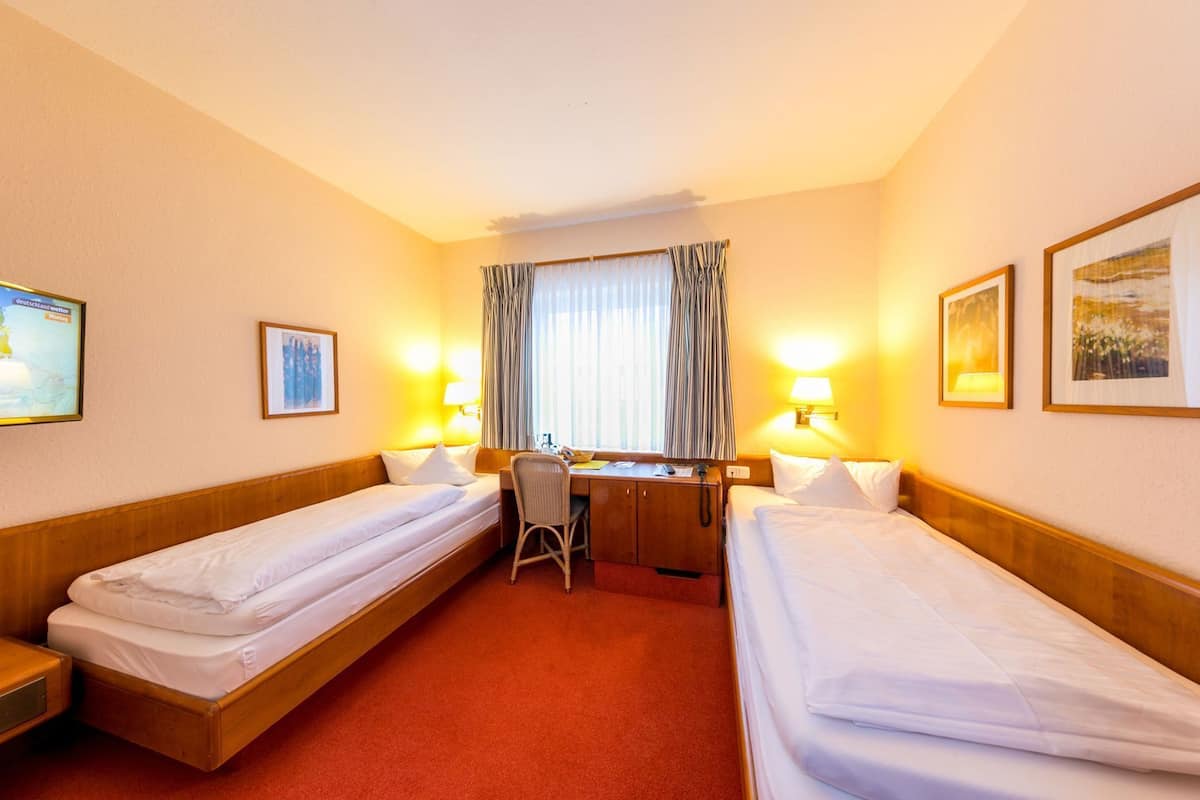 A hotel room with two single beds, a desk with a chair, a window with curtains, and two wall-mounted lamps.