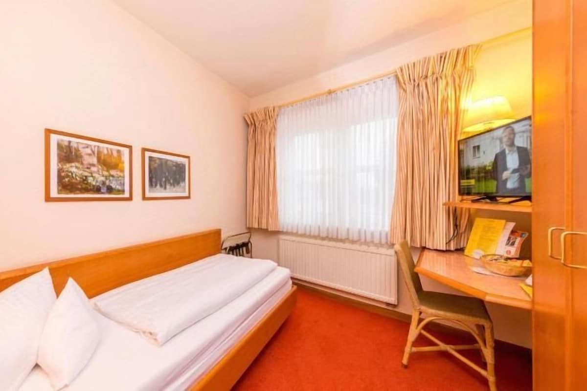 Une chambre d’hôtel avec un lit simple, un bureau avec une chaise, une télévision et deux tableaux encadrés au mur.