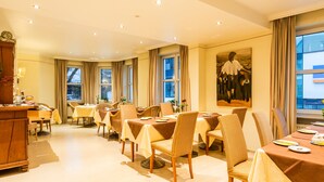 A dining area with multiple tables and chairs, large windows with curtains, and a painting on the wall. - CityHotel Kempten (Kempten)