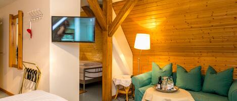 A cozy room with a wooden ceiling, a green sofa with pillows, a small table with a tray of bottles, a television mounted on the wall, and a bed with a white mattress.