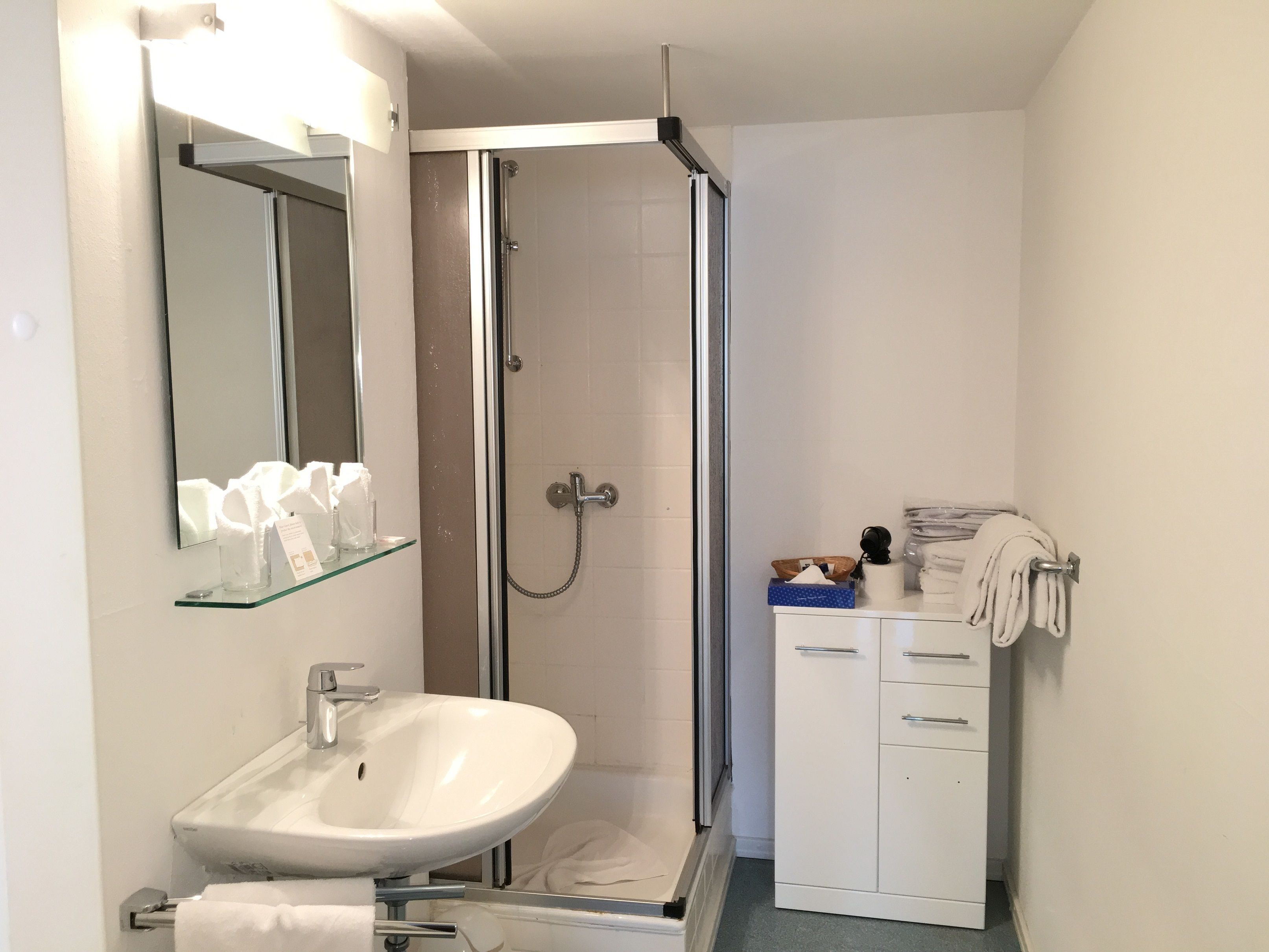 Ein Badezimmer mit Duschkabine aus Glas, einem weißen Waschbecken, einem Spiegel und einem Schrank mit Handtüchern.