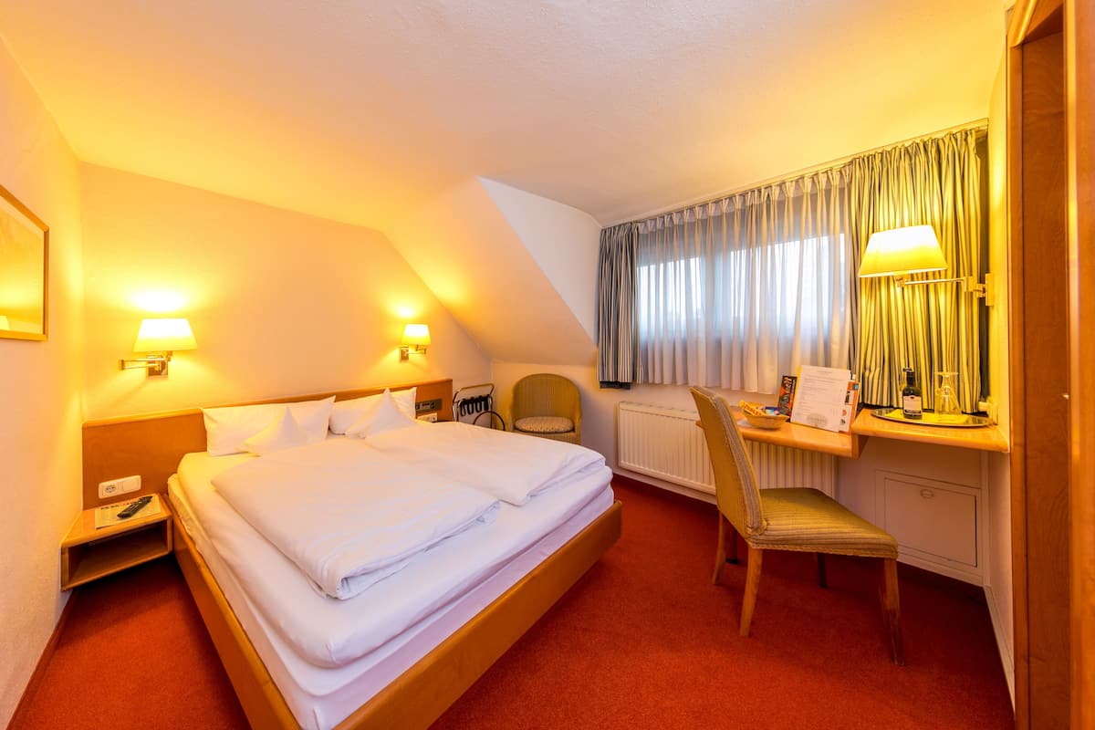 A hotel room with a bed, a desk with a lamp, a chair, and a window with curtains.