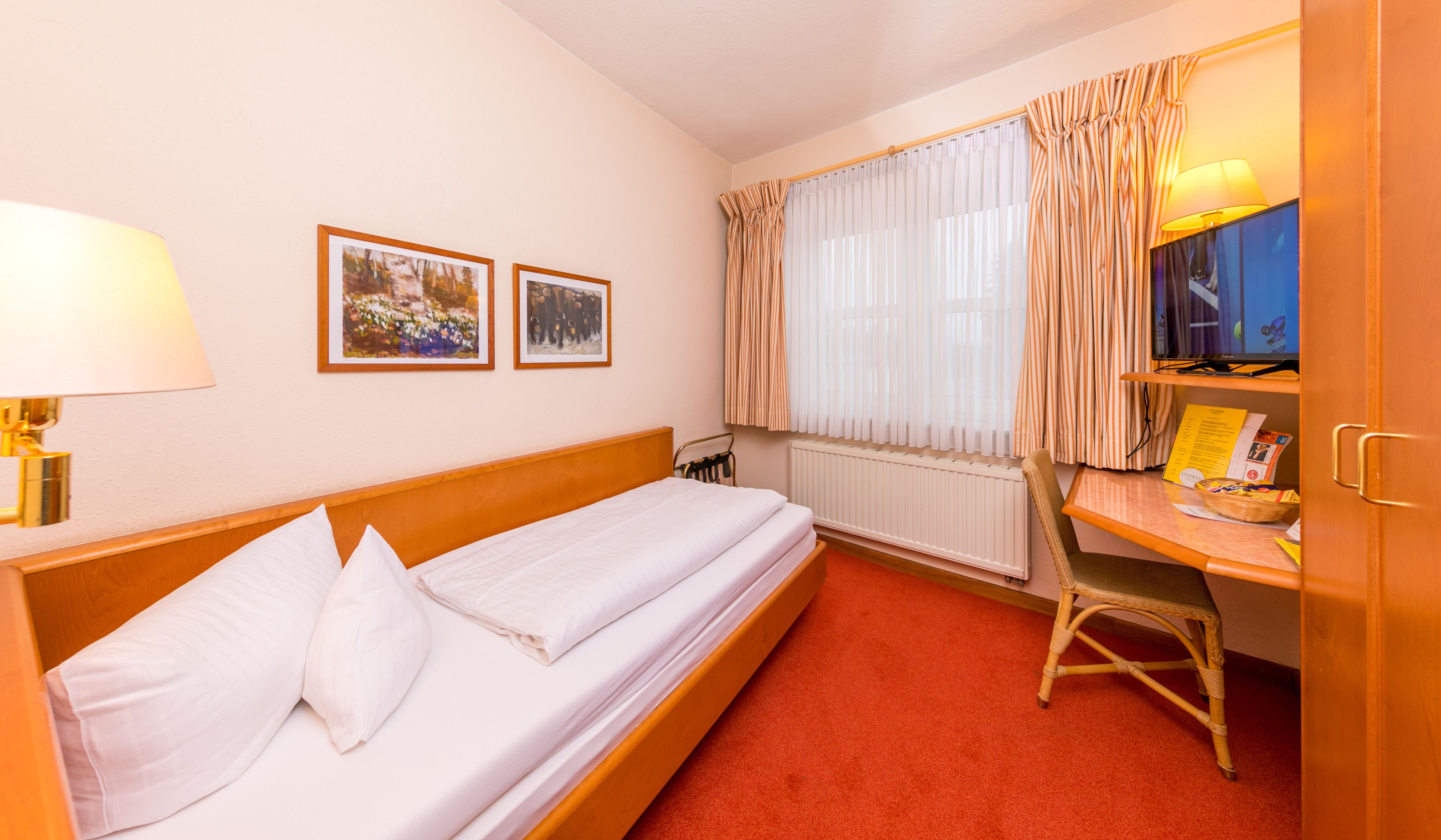 Ein Hotelzimmer mit einem Einzelbett, einem Schreibtisch mit Stuhl, einem Heizkörper und zwei gerahmten Bildern an der Wand.