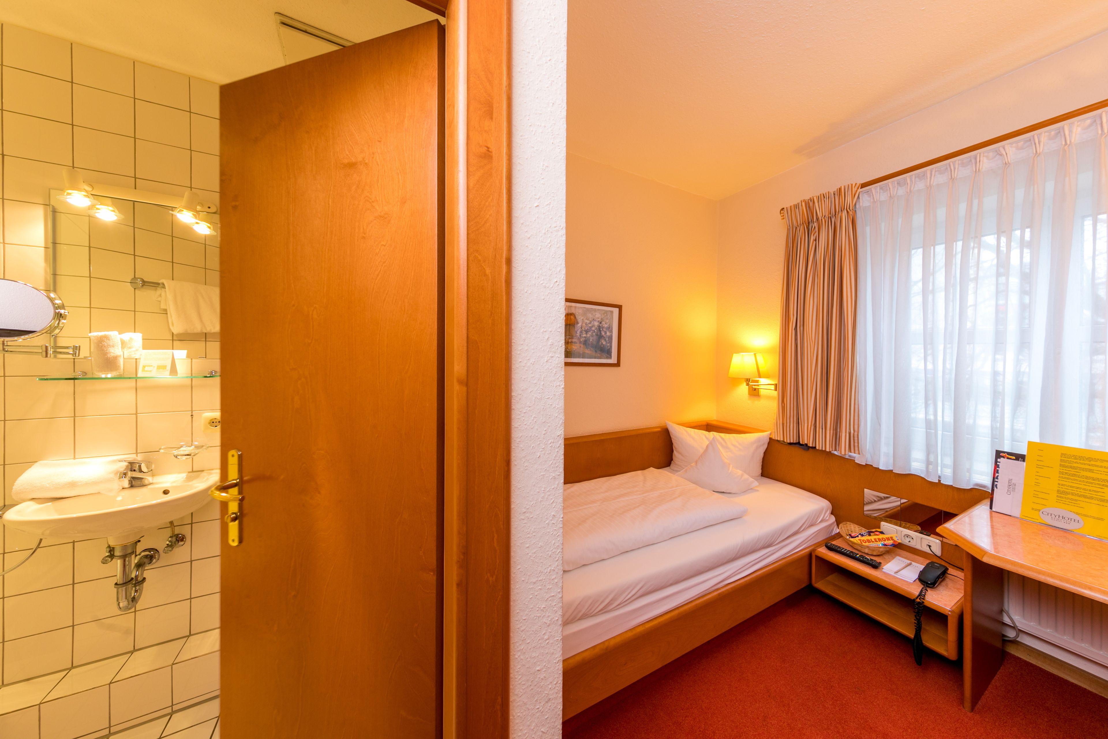 Habitación de hotel con puerta de madera, cama con ropa blanca, mesita de noche con lámpara, escritorio pequeño con silla y ventana con cortinas.