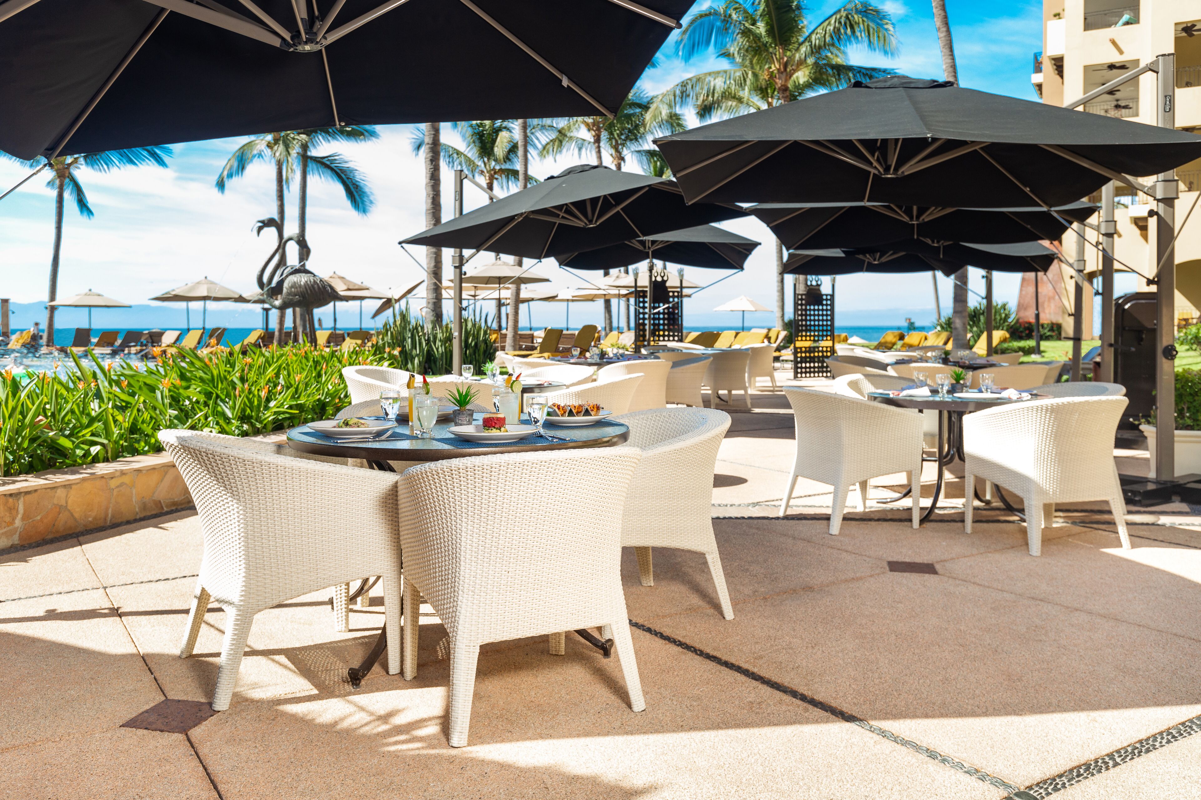 Een terras met witte rieten stoelen en tafels, grote parasols en uitzicht op de oceaan.