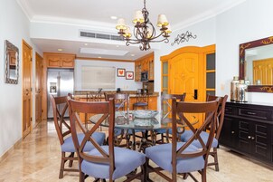 Un comedor con mesa de vidrio rodeada de sillas de madera, una lámpara de araña y armarios de madera.