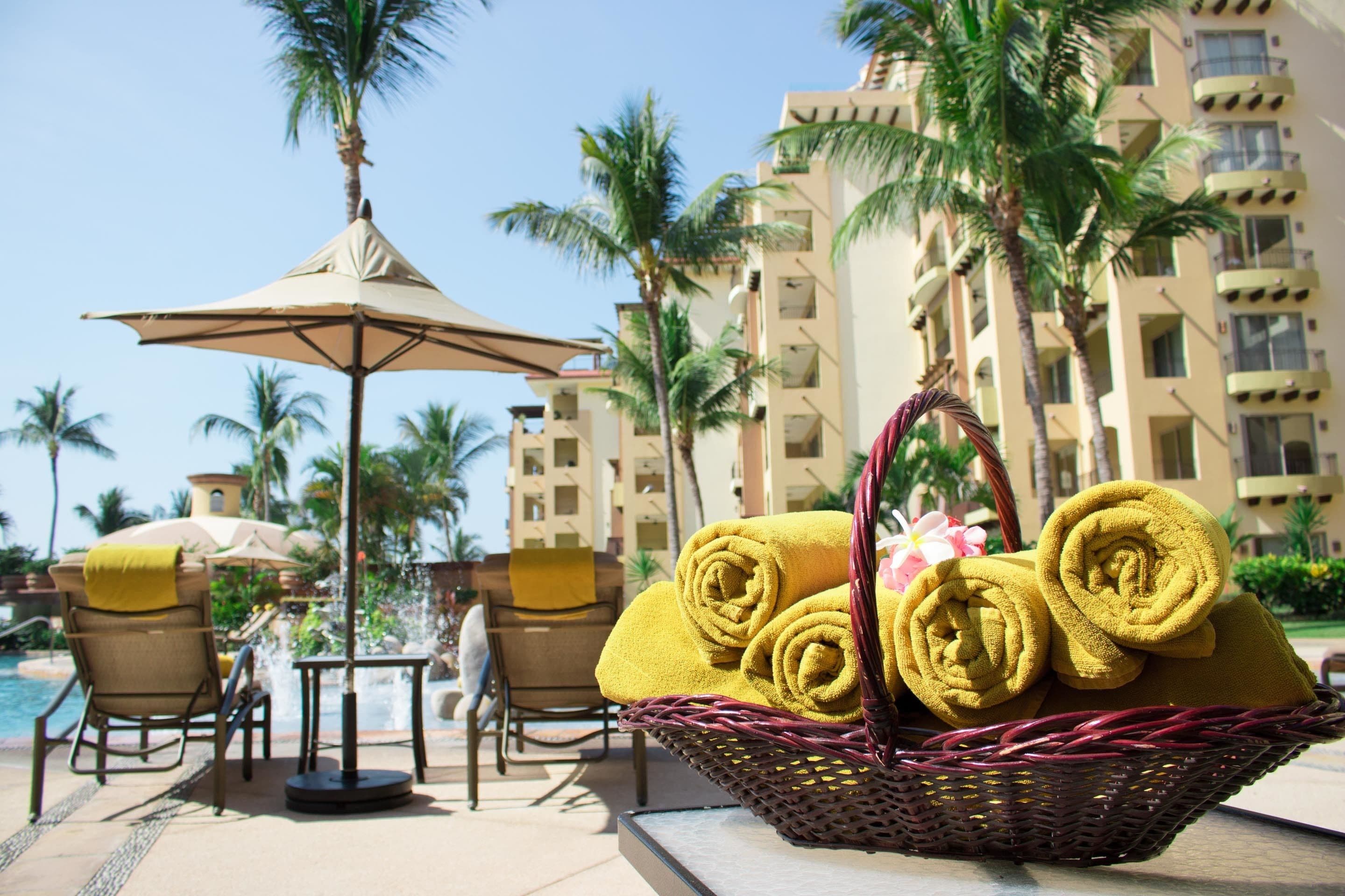 A poolside area with a wicker basket of rolled towels, lounge chairs, and a palm tree.