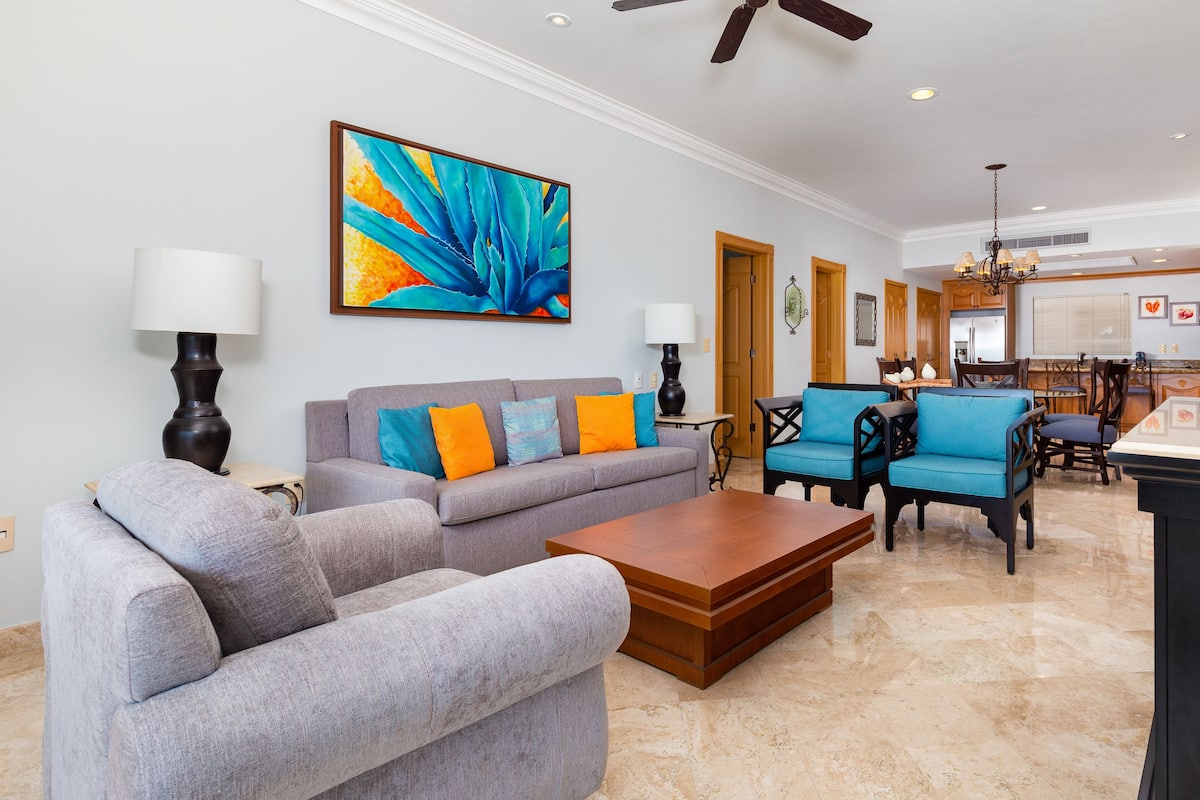 A spacious living room with a gray sofa, blue armchairs, and a wooden coffee table. A colorful abstract painting adorns the wall, and a ceiling fan is visible.