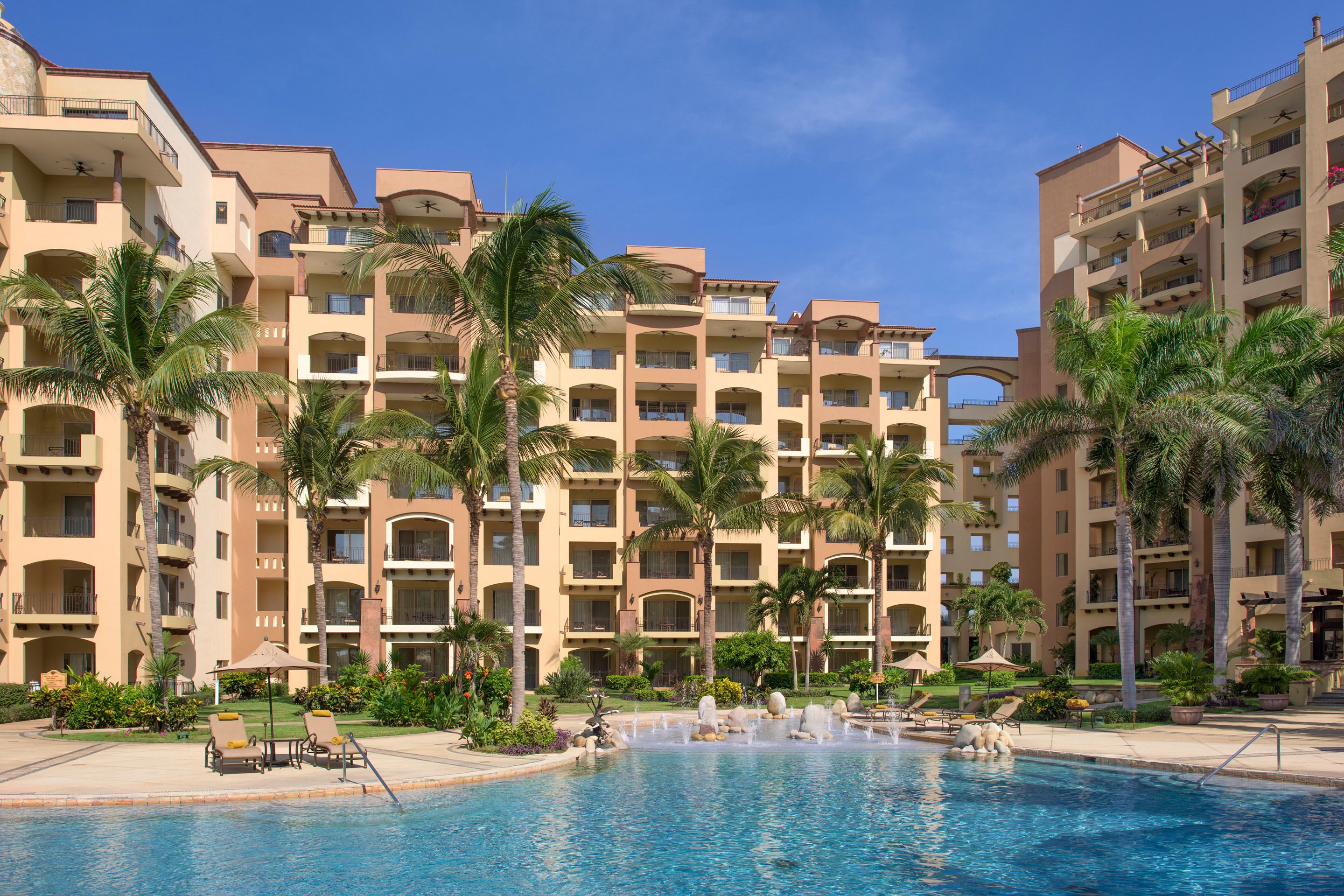 Un hôtel doté de nombreux balcons, d’une piscine avec une fontaine et de palmiers.
