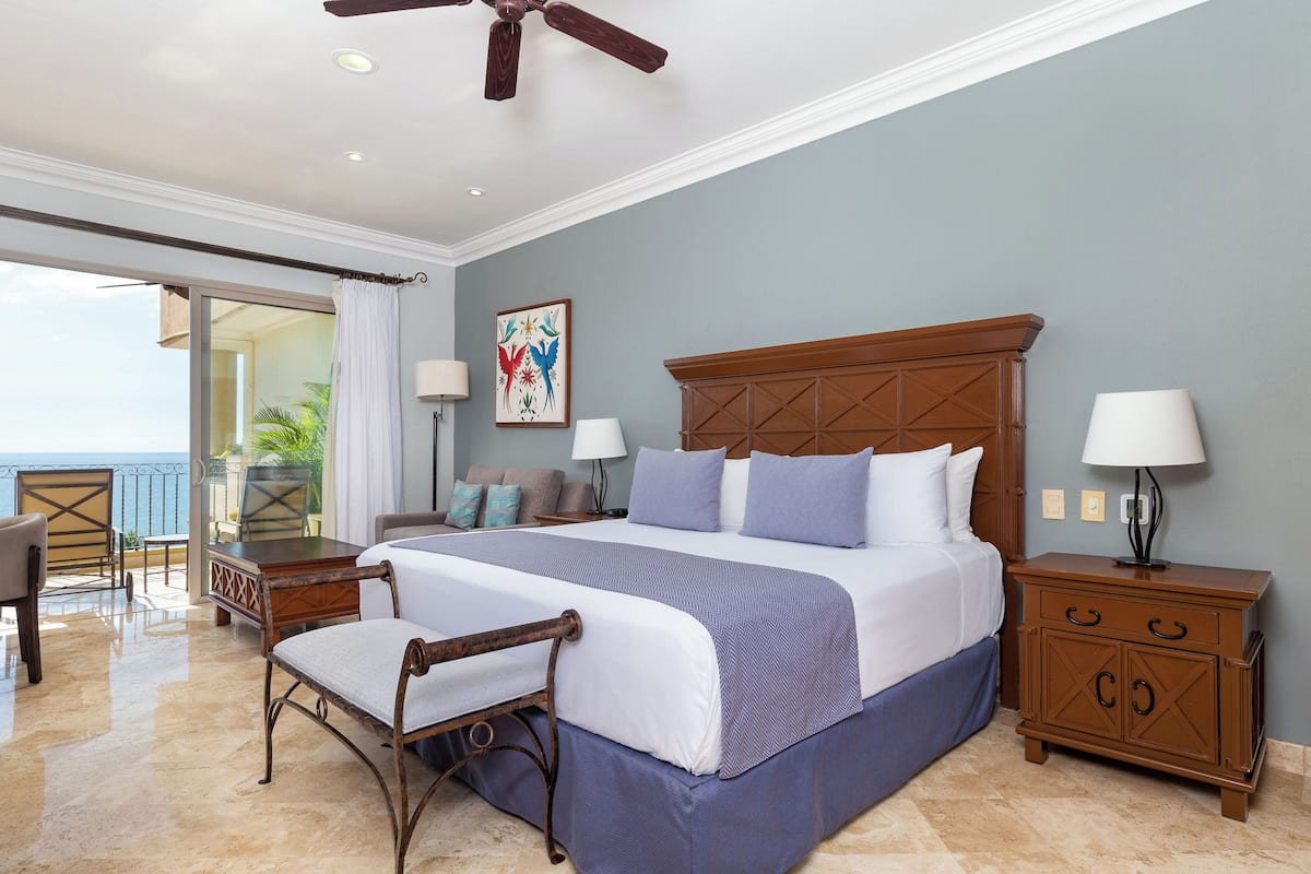 A bedroom with a large bed, a wooden nightstand, a chair, and a view of the ocean.