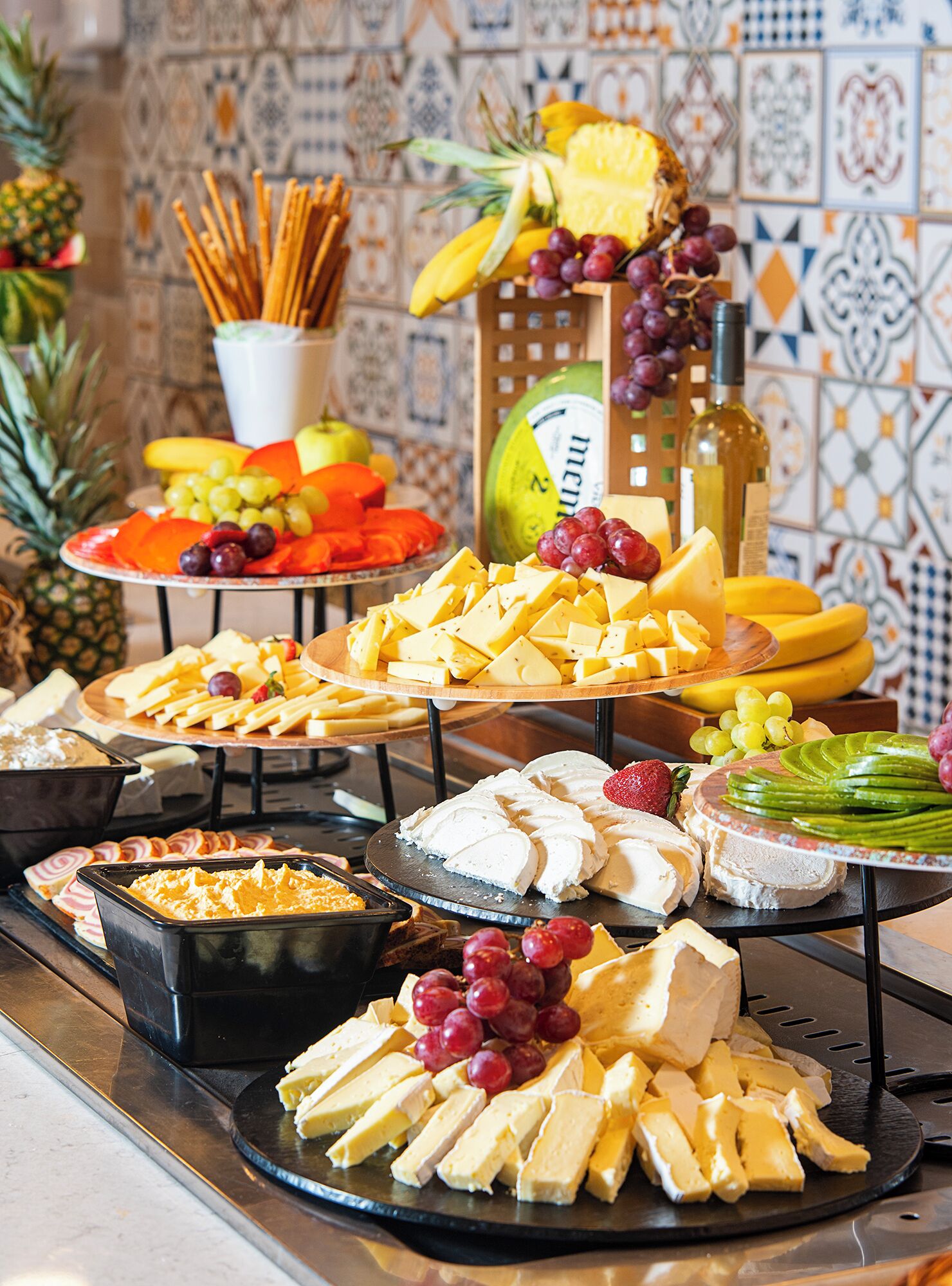 A buffet with various cheeses, fruits, and a bottle of wine.