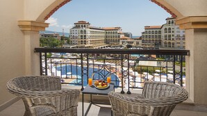 A balcony with wicker chairs, a table with fruit and drinks, and a view of a pool and buildings.