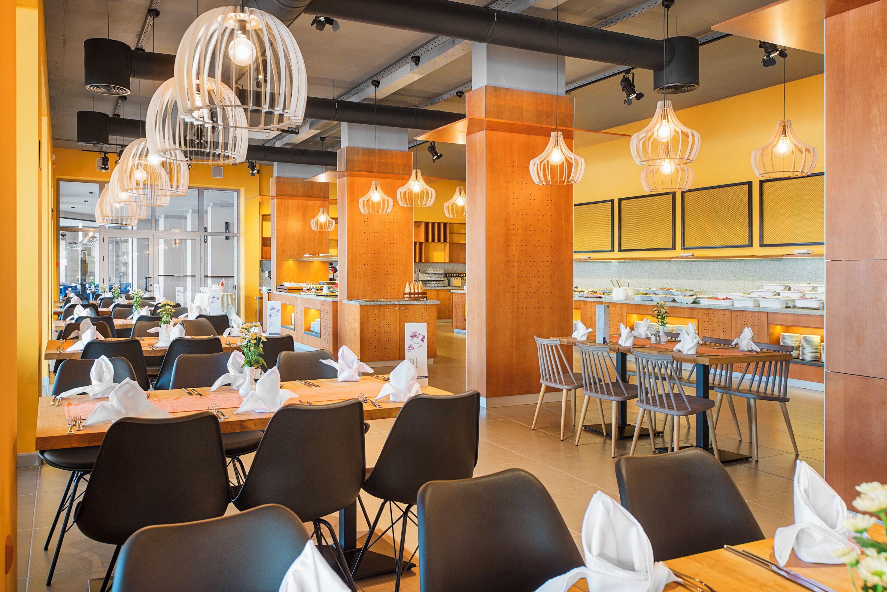 A modern dining area with multiple tables and chairs, overhead lighting, and a kitchen in the background.