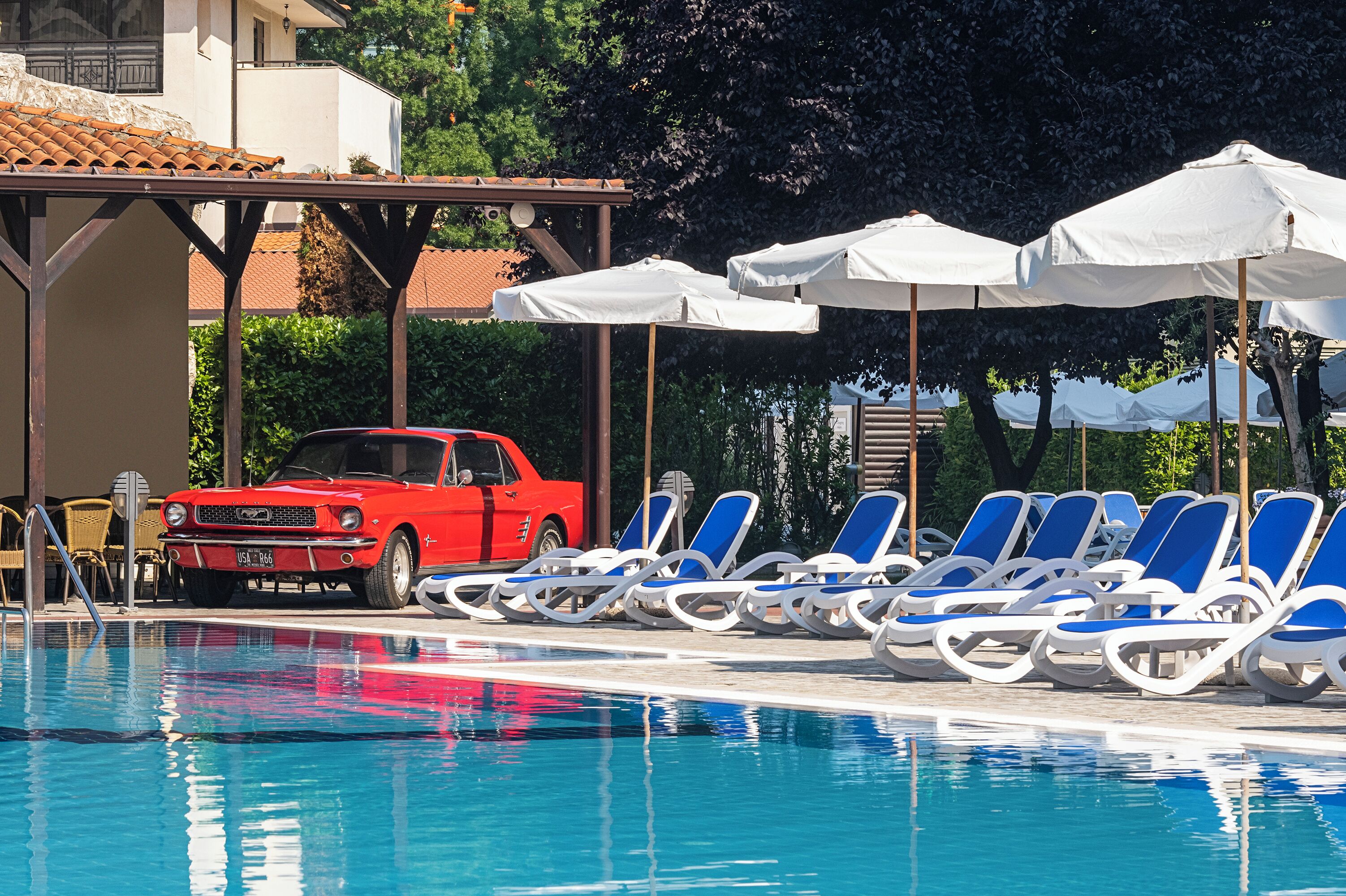 A swimming pool with blue lounge chairs and white umbrellas, a red car, and a building with a tiled roof in the background.