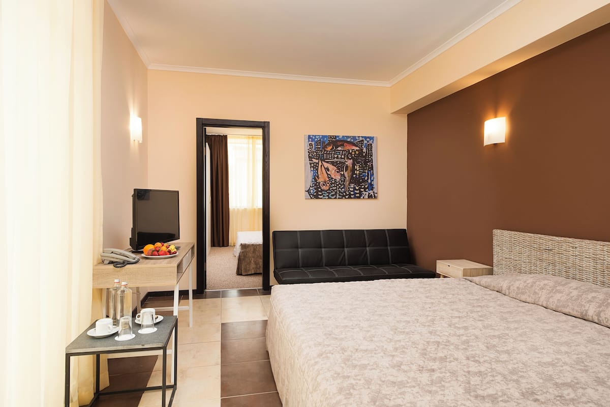 A hotel room with a bed, a desk with a phone and a plate of fruit, a television, and a painting on the wall.