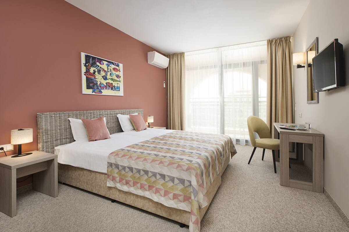 A hotel room with a bed, a desk, a chair, a TV, and a window with curtains.