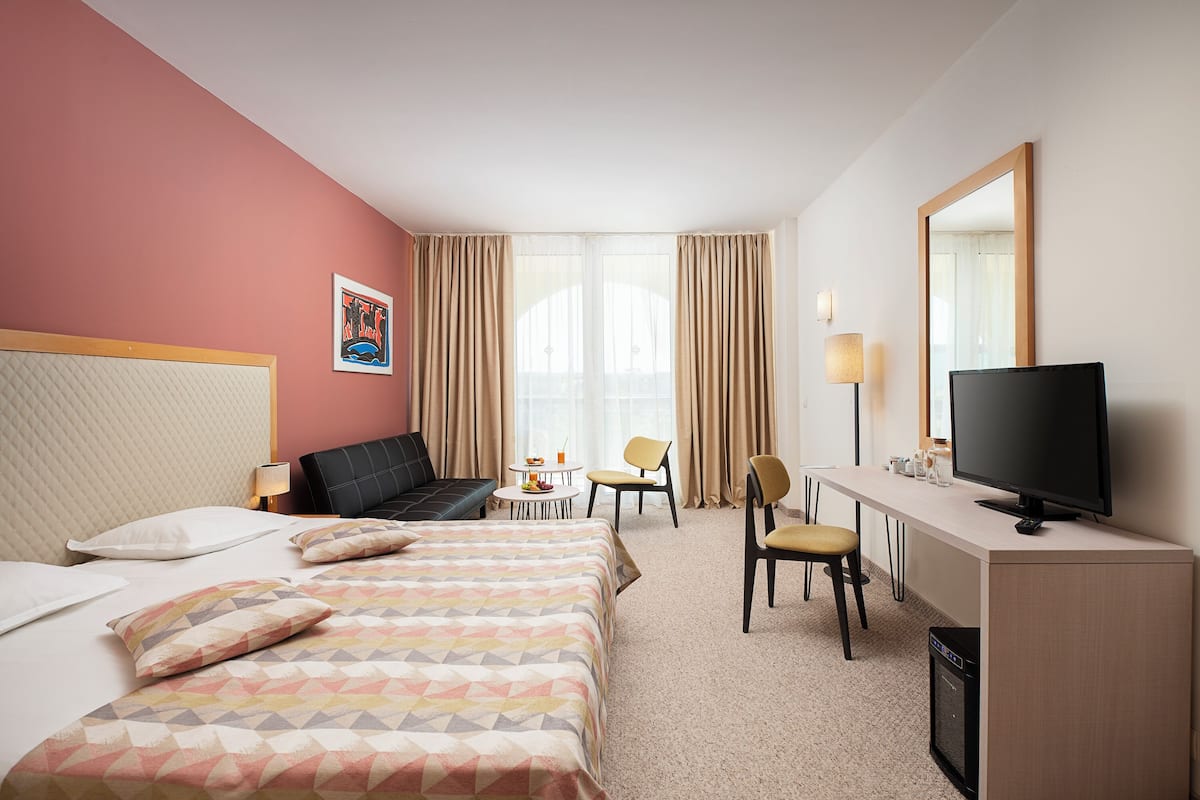A hotel room with a bed, a desk with a television, a chair, a small table with a lamp, and a window with curtains.