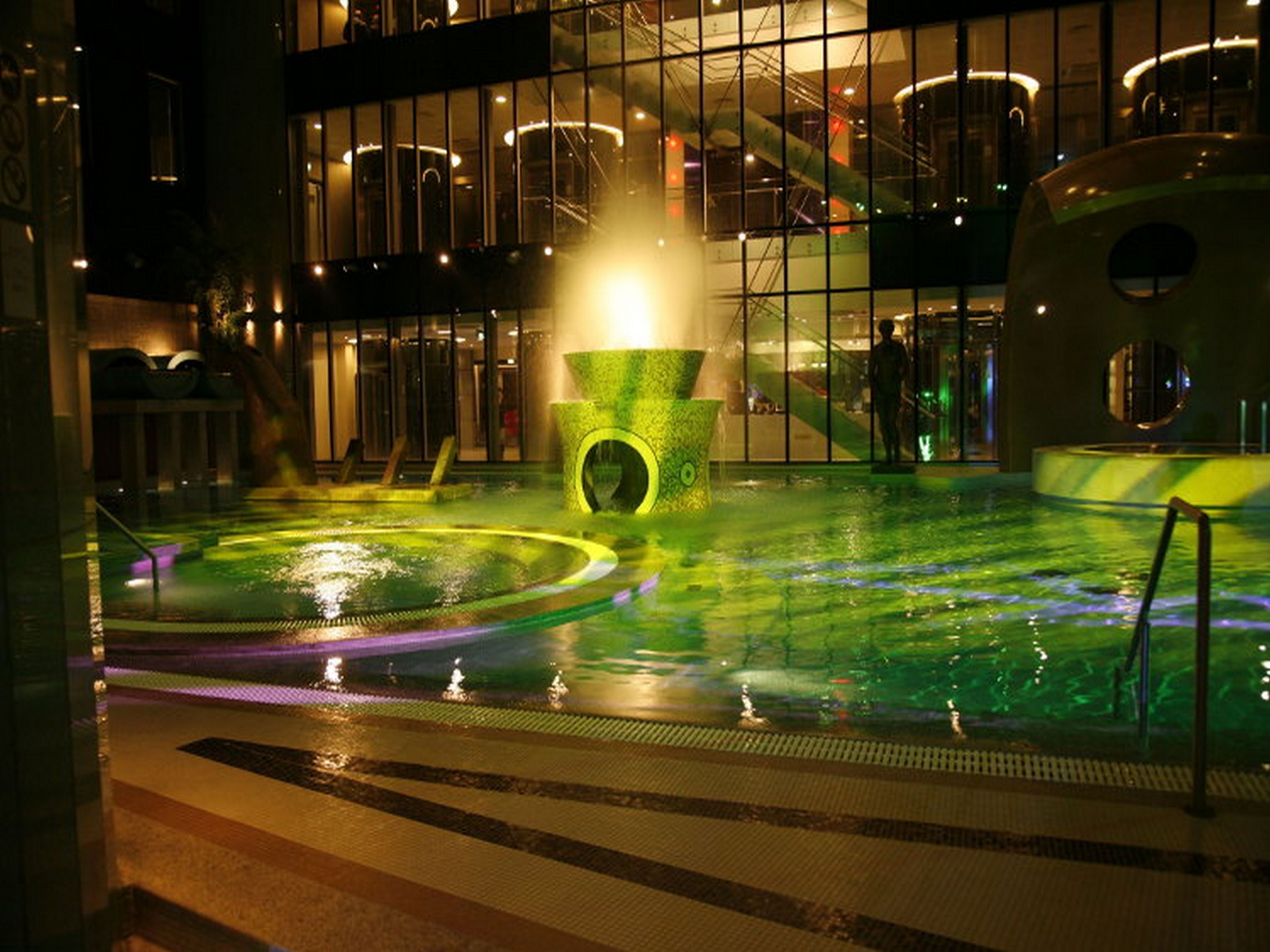 An indoor pool area with a central water feature, surrounded by a glass wall reflecting the interior lights.