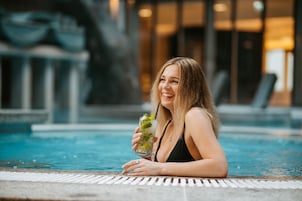 A person sitting by a pool with a drink, surrounded by a modern building with glass windows.