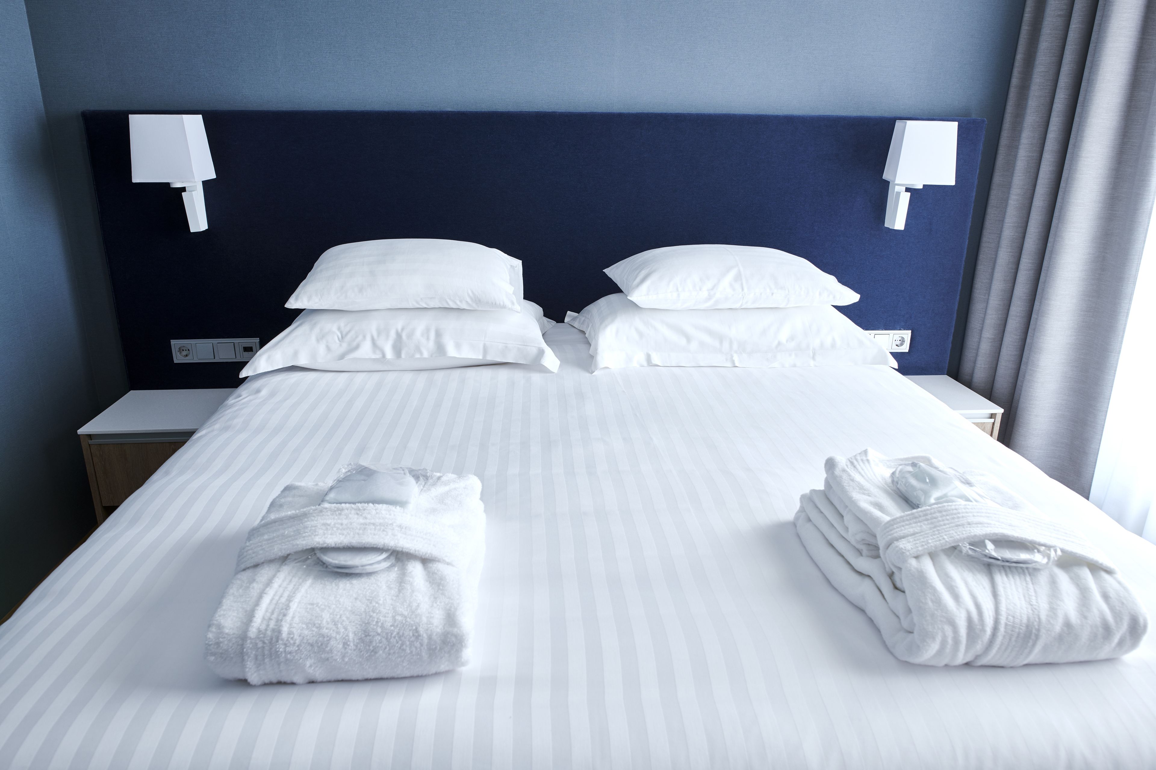 A hotel room with a neatly made bed, two white towels, a nightstand, and a wall-mounted light fixture.