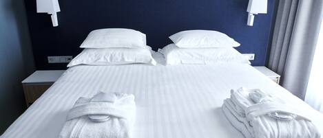 A hotel room with a neatly made bed, two white towels, a nightstand, and a wall-mounted light fixture.
