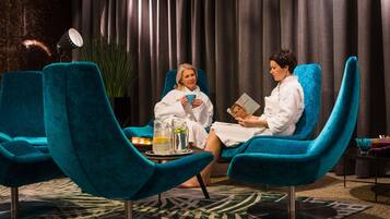 Two individuals seated in a teal-colored lounge chair in a room with a patterned rug, a small table with a pitcher and glasses, and a lamp in the background.