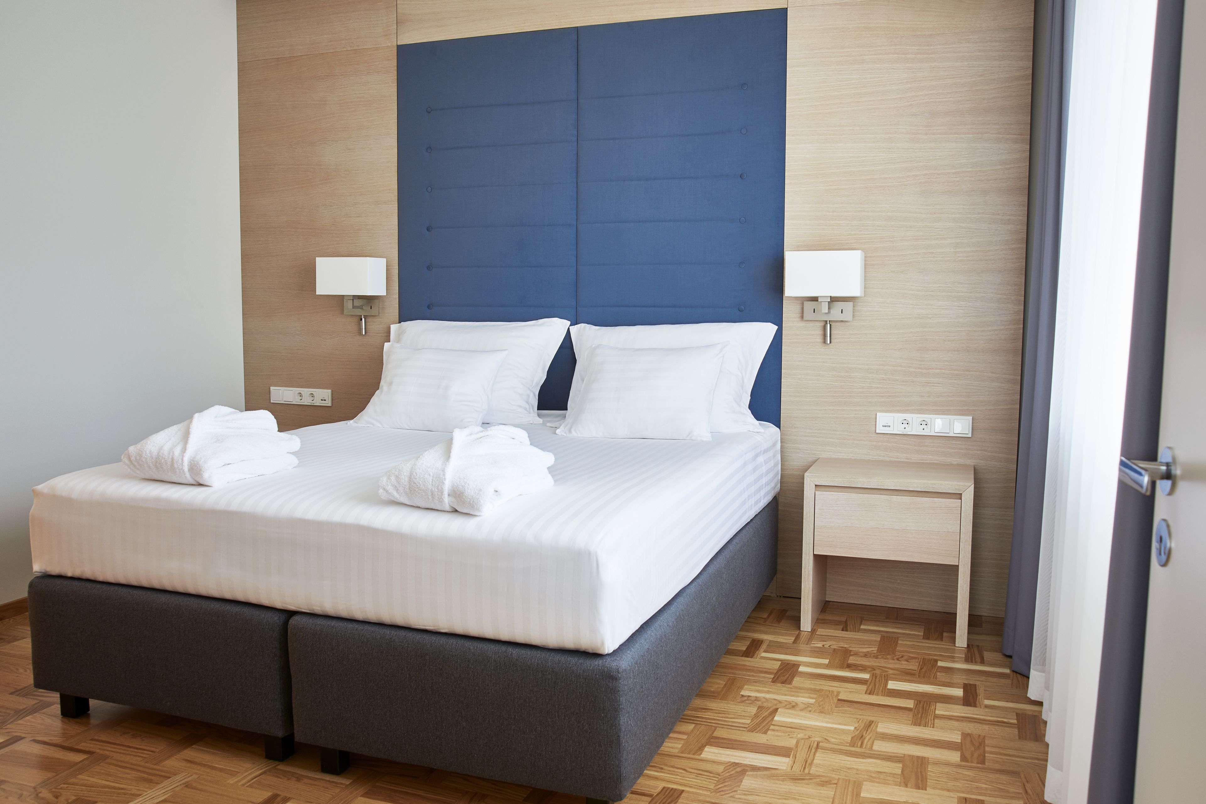 A neatly made bed with white linens and two pillows, a bedside table with a lamp, and a wooden floor.