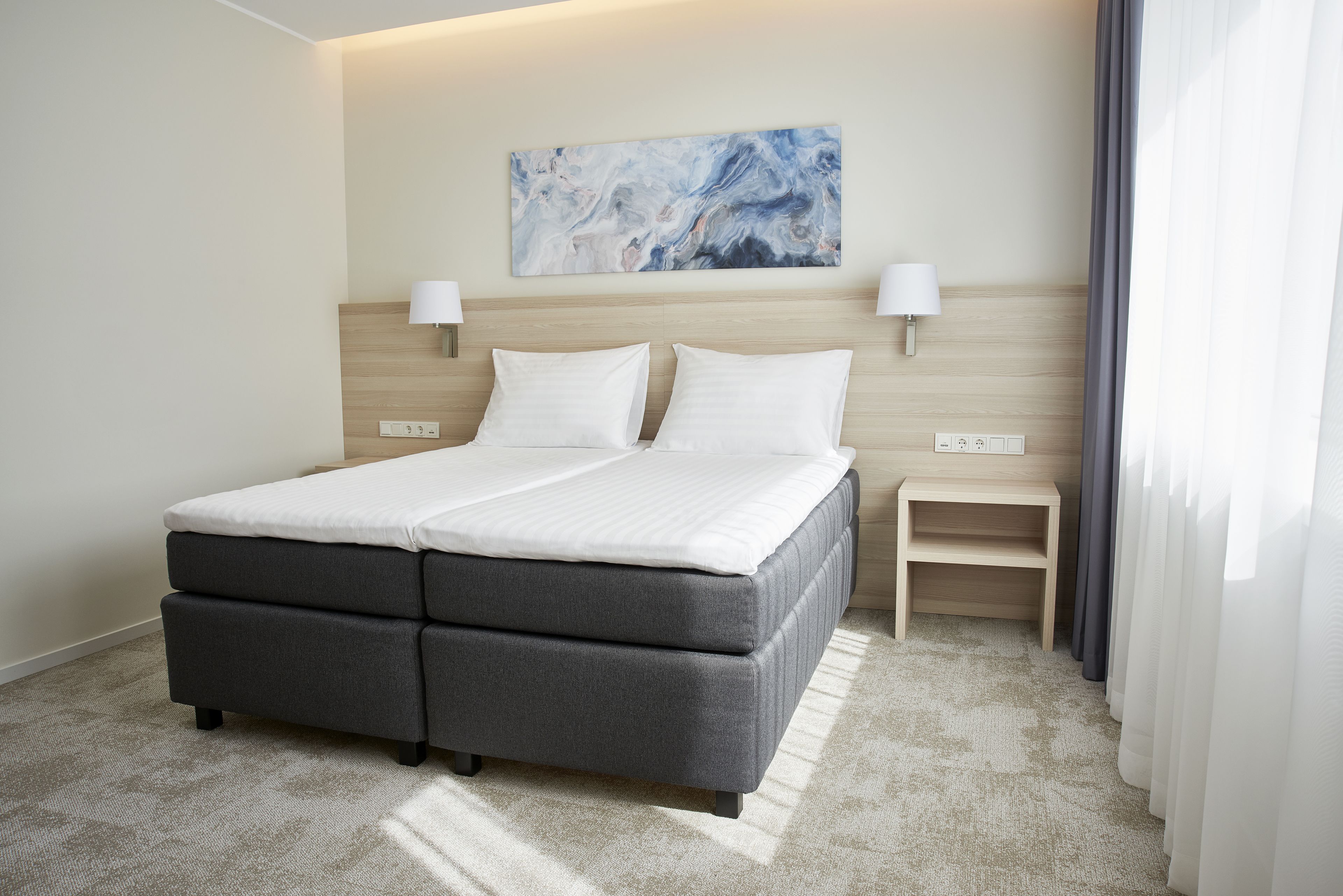 Una cama doble con cabecera tapizada de gris, dos mesitas de noche con lámparas, una cabecera de color claro y una obra de arte con diseño de mármol sobre la cama.