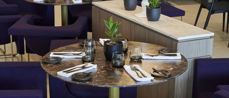 Un comedor moderno con mesas redondas, sillas moradas y una isla central con una lámpara y plantas.