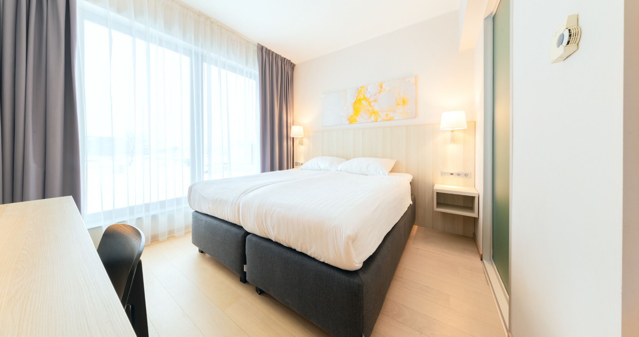 A modern hotel room with a large bed, a desk, and a chair. There is a window with curtains, a wall-mounted light, and a small shelf with a lamp.