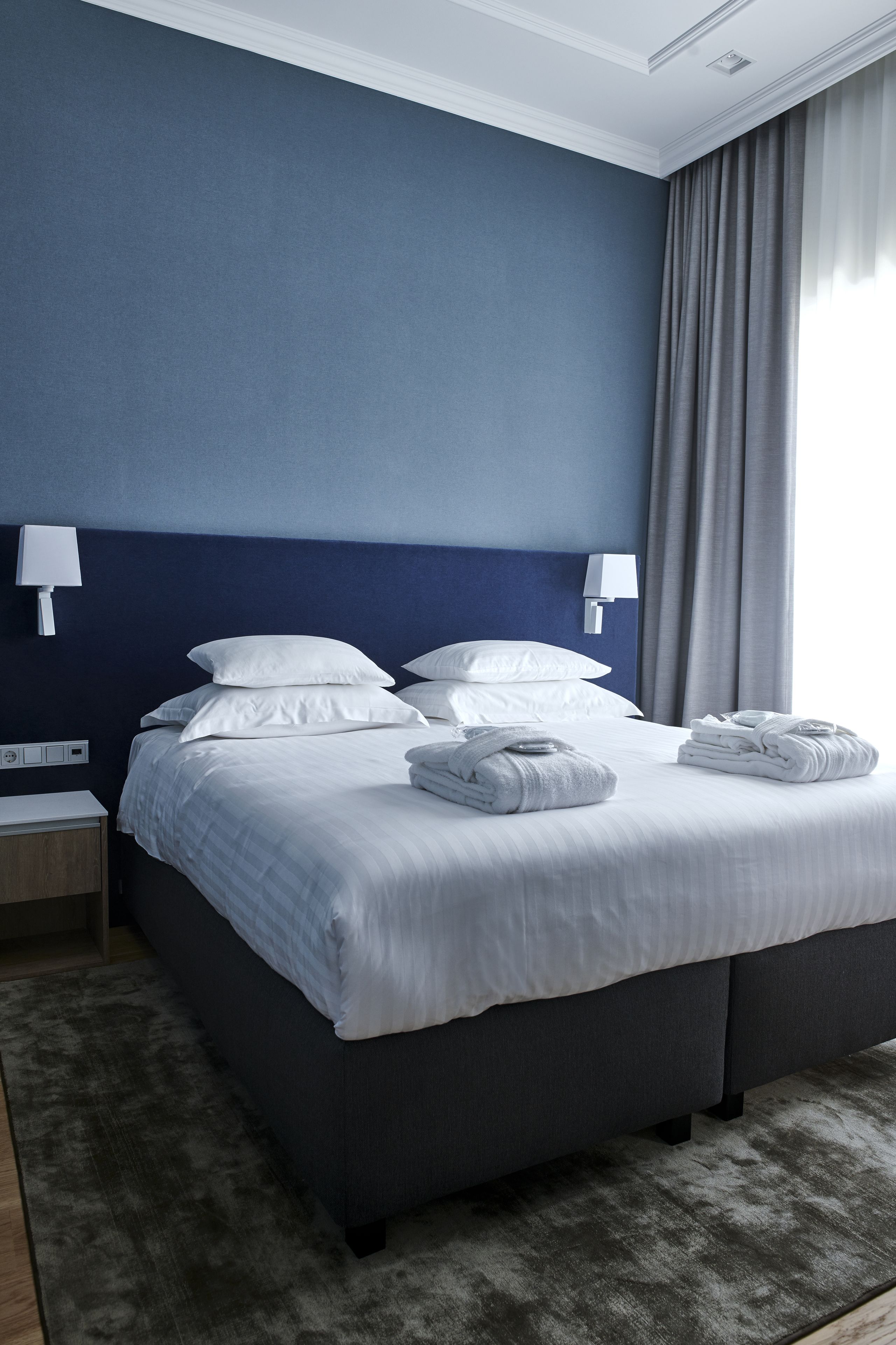 A hotel room with a large bed, white linens, a dark blue headboard, a nightstand with a lamp, and a window with curtains.