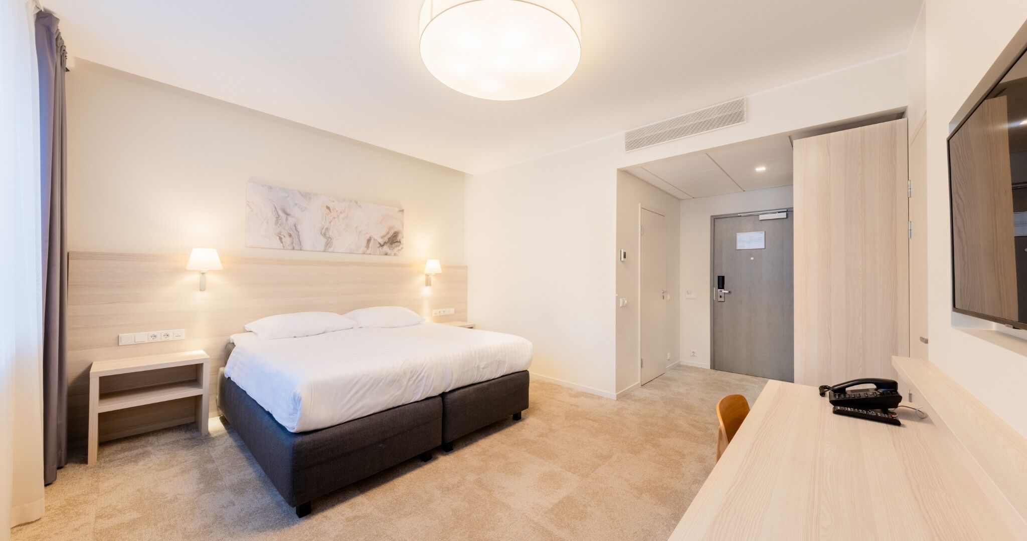 Une chambre d’hôtel moderne équipée d’un lit, de tables de chevet, d’un bureau et d’un grand miroir.