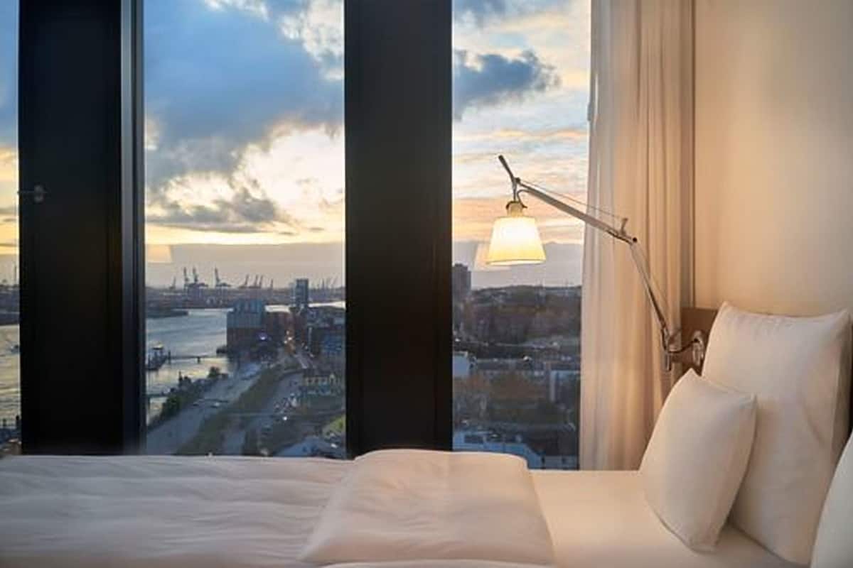 A hotel room with a large bed, a bedside lamp, and a view of a cityscape through large windows.