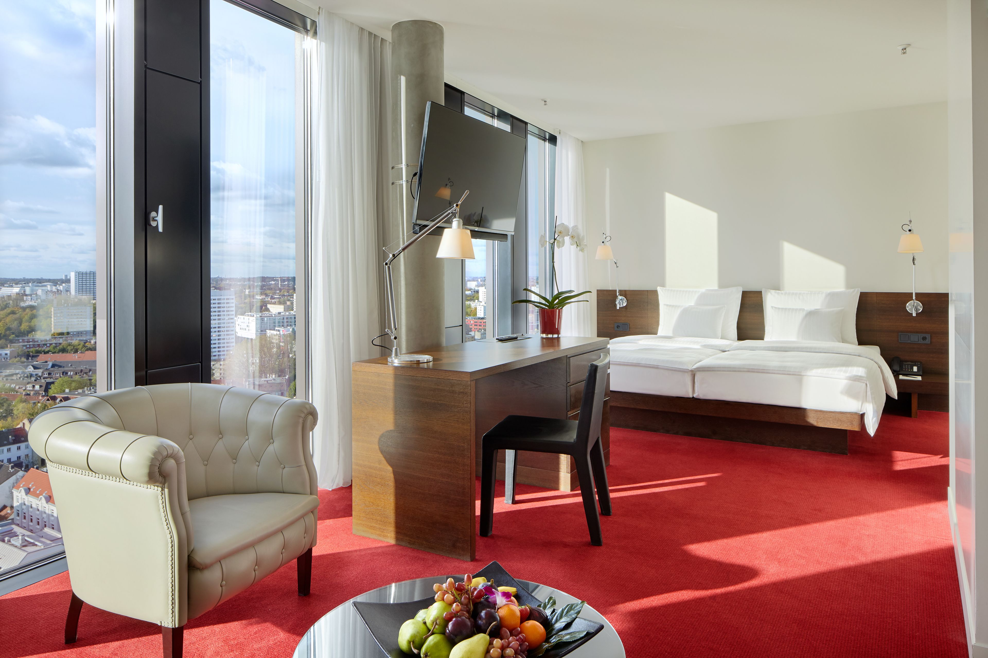 A hotel room with a large bed, a desk, a chair, a red carpet, a view of the city, and a window with curtains.