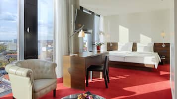 A hotel room with a large bed, a desk, a chair, a red carpet, a view of the city, and a window with curtains.