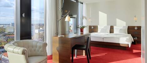 A hotel room with a large bed, a desk, a chair, a red carpet, a view of the city, and a window with curtains.