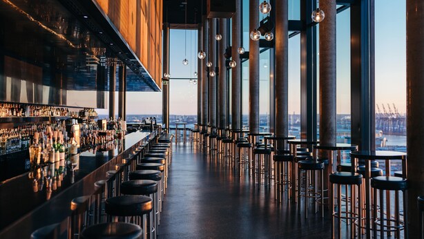 A bar with a long counter and stools, large windows, and a view of the city. - Empire Riverside Hotel (Hamburg)