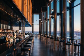 Un bar con una barra larga y taburetes, ventanas amplias y vistas a la ciudad.