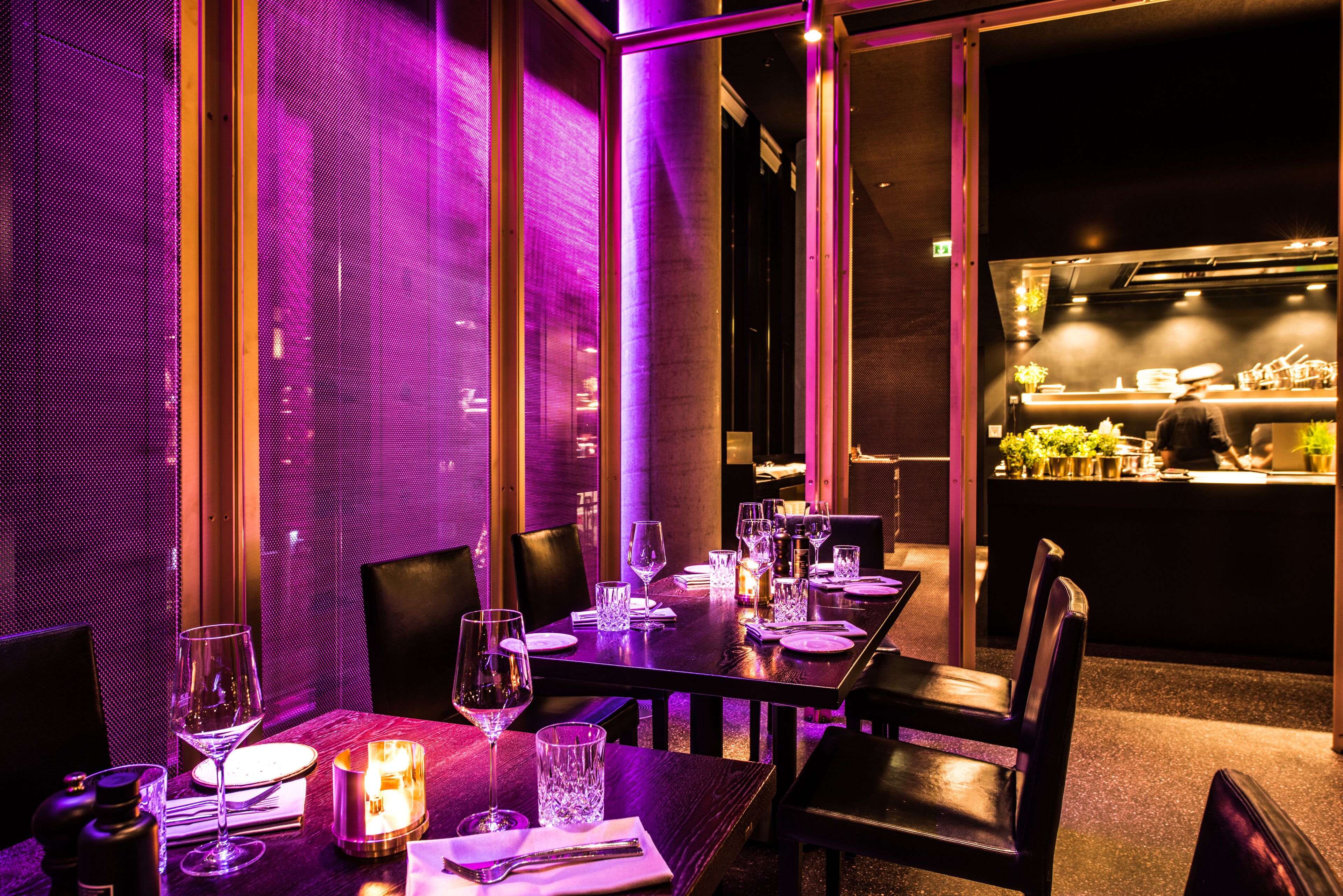 Un restaurante con mesas dispuestas para comer, una zona de bar al fondo e iluminación ambiental morada.