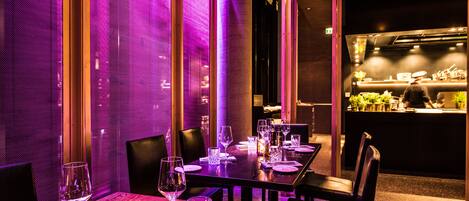 Un restaurante con mesas dispuestas para comer, una zona de bar al fondo e iluminación ambiental morada.